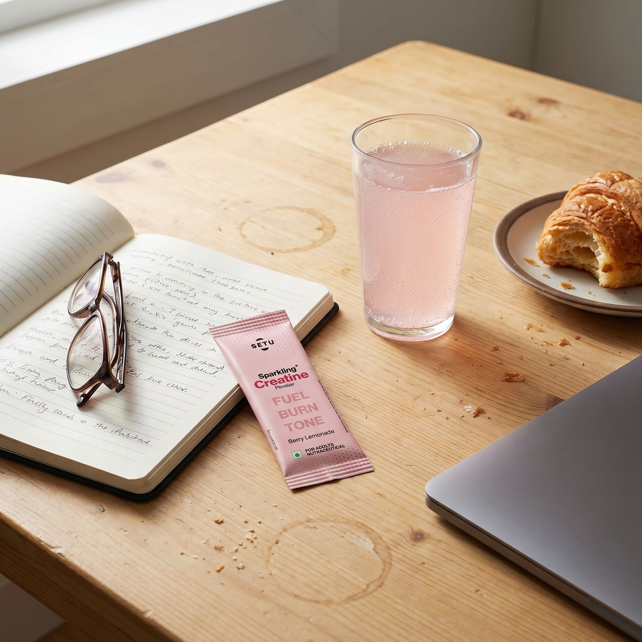 A real wood morning desk: SETU Sparkling Creatine sachet beside an open notebook with handwritten notes, folded reading glasses, a tall glass of fizzing pale-pink berry-lemonade, a half-eaten croissant on a plate, and the edge of a closed laptop